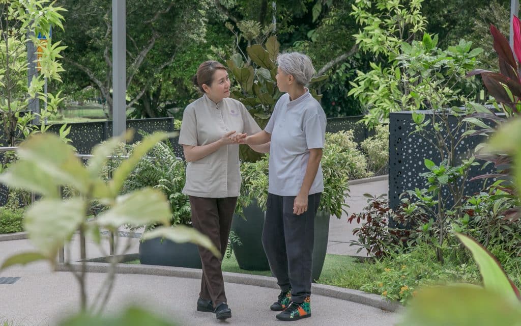 Elderly person walking with support along a garden path, surrounded by lush greenery